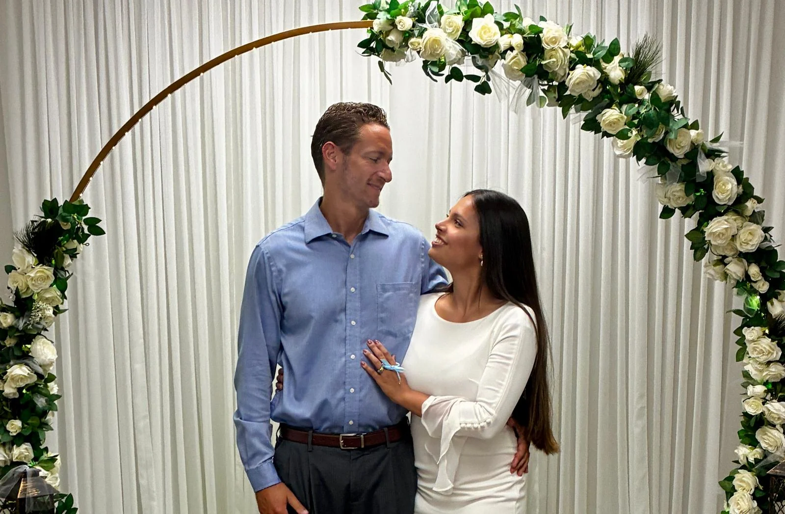 Rob and Veronica smiling at each other beneath a white floral arch